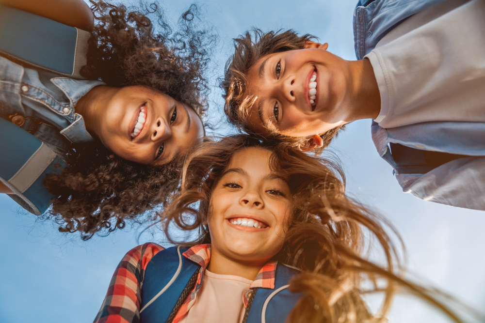A group of three happy, diverse children huddle together, smiling down at the camera. Give your child a healthy, confident smile with Pediatric Dentistry at Wilder Dentistry