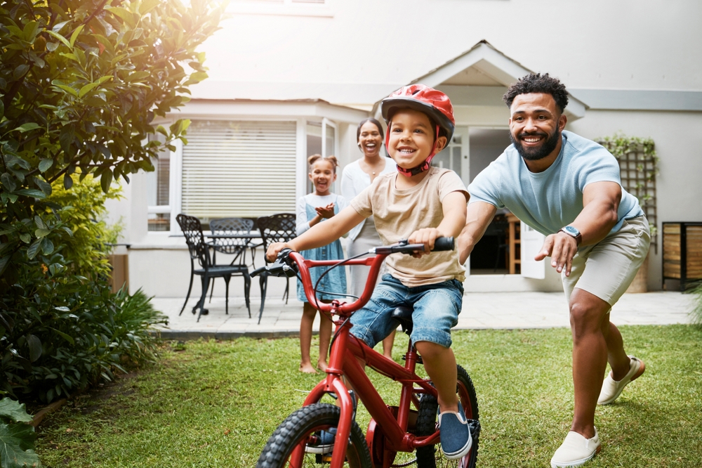 A joyful boy with a bright smile learns to ride his bike in the backyard with his father, as his mother and sister cheer him on. A healthy, active childhood is supported by a great Oral Care Routine For Children in Spokane Valley