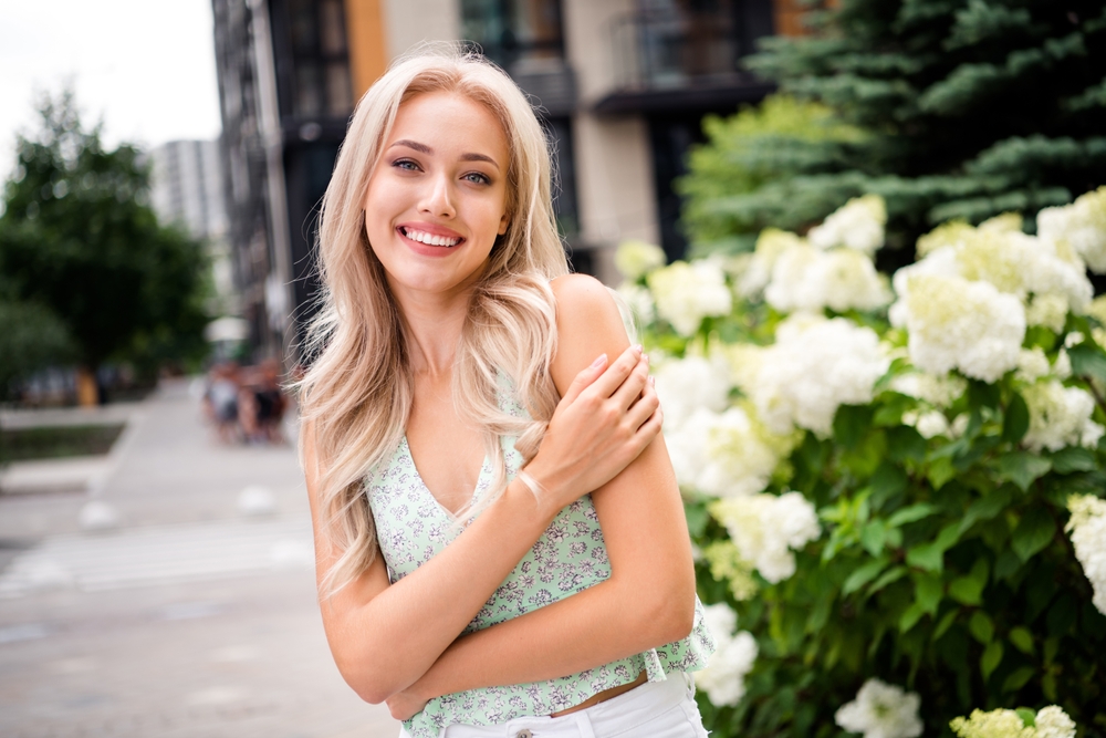 A young woman with a stunningly bright, white smile stands outdoors next to a flowering bush. Get a radiant smile like hers with professional Teeth Whitening at Wilder Dentistry