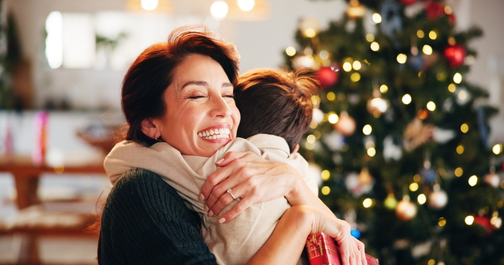 A mother with a radiant smile joyfully hugs her child by a Christmas tree. Don't miss a single precious moment—restore your smile quickly with Same-Day Crowns at Wilder Dentistry
