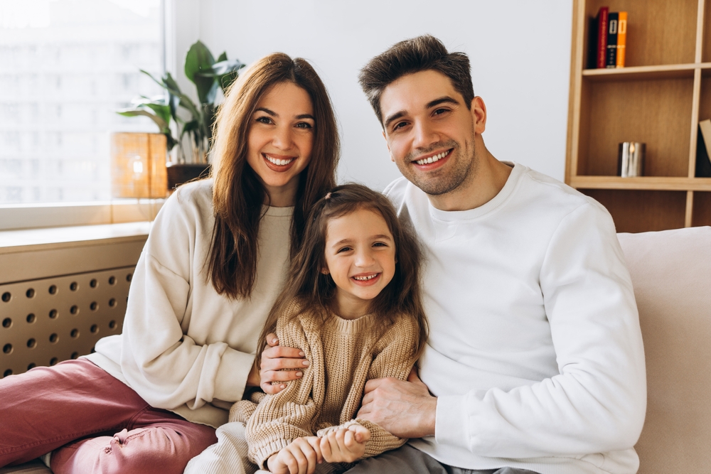 A happy family—mother, father, and young daughter—sit together on a couch, all sharing bright, healthy smiles thanks to comprehensive family Dental Services in Spokane Valley
